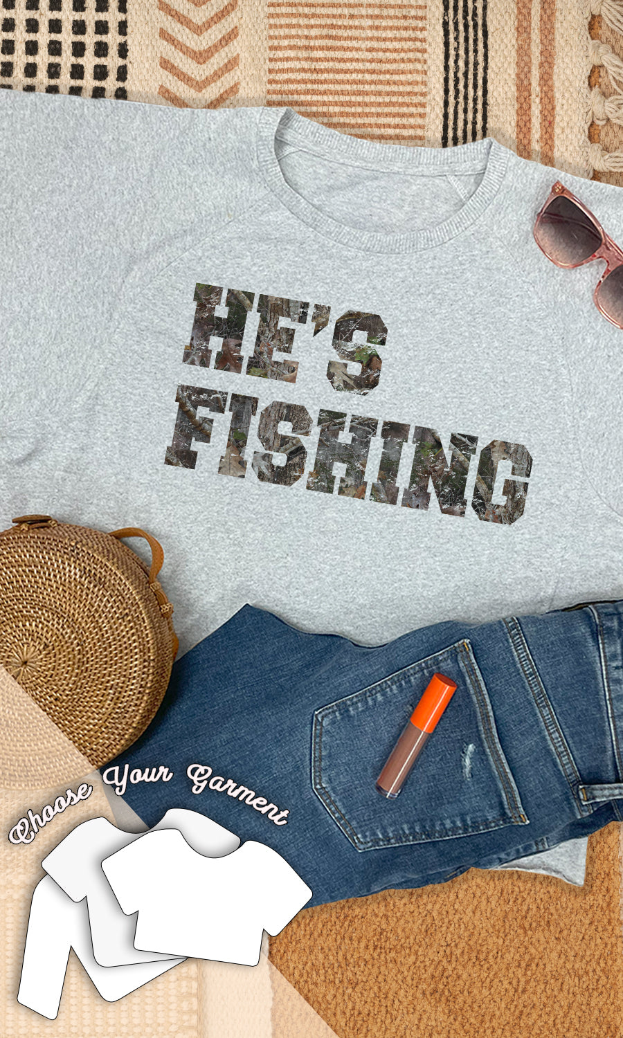 A graphic t-shirt with the text 'HE'S FISHING' printed on it, displayed flat with a pair of jeans, a pair of sunglasses, and a fishing reel.